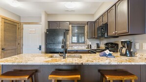 A kitchen with a granite countertop, stainless steel appliances, wooden bar stools, and dark cabinetry. - Walk In Condo just 2 Min to SDC, Resort Perks, Lake Access, Ticket Deals! (Branson)