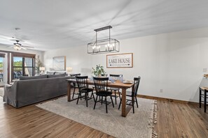 Un salon moderne avec un canapé gris, une table à manger avec des chaises, un lustre et un panneau d'accueil encadré.