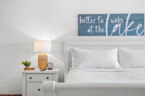 Ein Schlafzimmer mit einem weißen Bett, einem Nachttisch mit Lampe, einer Uhr und einem dekorativen Schild an der Wand.