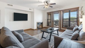 A modern living room with a flat-screen TV, a fireplace, a ceiling fan, and a large window with blinds. - 2 bd/2ba, Deck Space, Ticket Deals and 2 minutes to Silver Dollar City! (Branson)