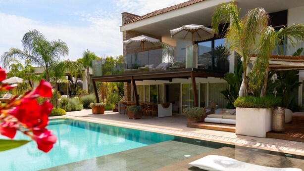 Une piscine avec des chaises longues, un patio agrémenté de parasols et une maison dotée d’un balcon donnant sur la piscine.