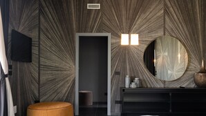 Une chambre moderne avec un grand lit, un miroir rond, un mur en bois foncé orné d’un motif radial, un petit pouf rond et une console noire sur laquelle est posé un vase.