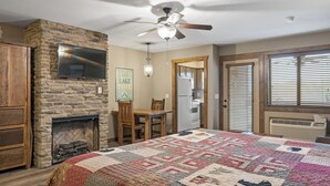 Una habitación acogedora con chimenea de piedra, colcha acolchada, ventilador de techo y televisor montado en la pared.