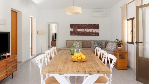 A dining area with a wooden table, white chairs, a television, and a painting on the wall. - Holiday Home 'Ca N'Alicia Des Bosch' with Mountain Views, Private Terrace and Wi-Fi (Illes Balears)