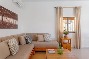 A living room with a sofa, cushions, a small table with a bottle, a plant, and a window with curtains.