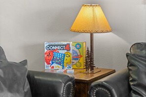 A Connect 4 game box, a lamp, and a small tree model on a wooden side table in a room with leather chairs.