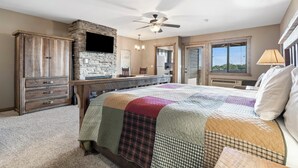 A bedroom with a stone fireplace, a ceiling fan, a large bed with a quilt, a wooden dresser, and a window with blinds. - Studio Condo overlooking Table Rock Lake, 2 min to SDC, Ticket Deals, Resort Perks! (Branson)