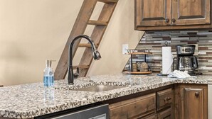Eine Küche mit einer Granit-Arbeitsplatte, einer Edelstahl-Spüle, Holzschränken, einer Kaffeemaschine und einer Leiter.