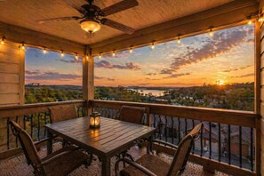 Eine Holzterrasse mit Tisch und Stühlen, einem Deckenventilator, Lichterketten und Blick auf einen Sonnenuntergang über der Landschaft.