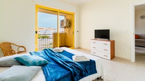 Une chambre à coucher avec un lit, un téléviseur posé sur une commode en bois, un fauteuil en osier et une porte-fenêtre coulissante donnant sur un balcon.