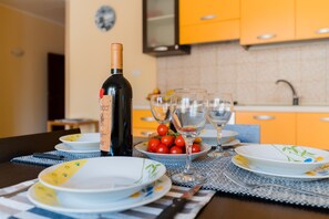 Une table à manger est dressée avec des assiettes, une bouteille de vin et des verres, et un saladier de tomates cerises est posé à l’arrière-plan.