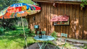 Un bâtiment rustique en bois, avec une petite table et deux chaises, un parasol et une bouteille.