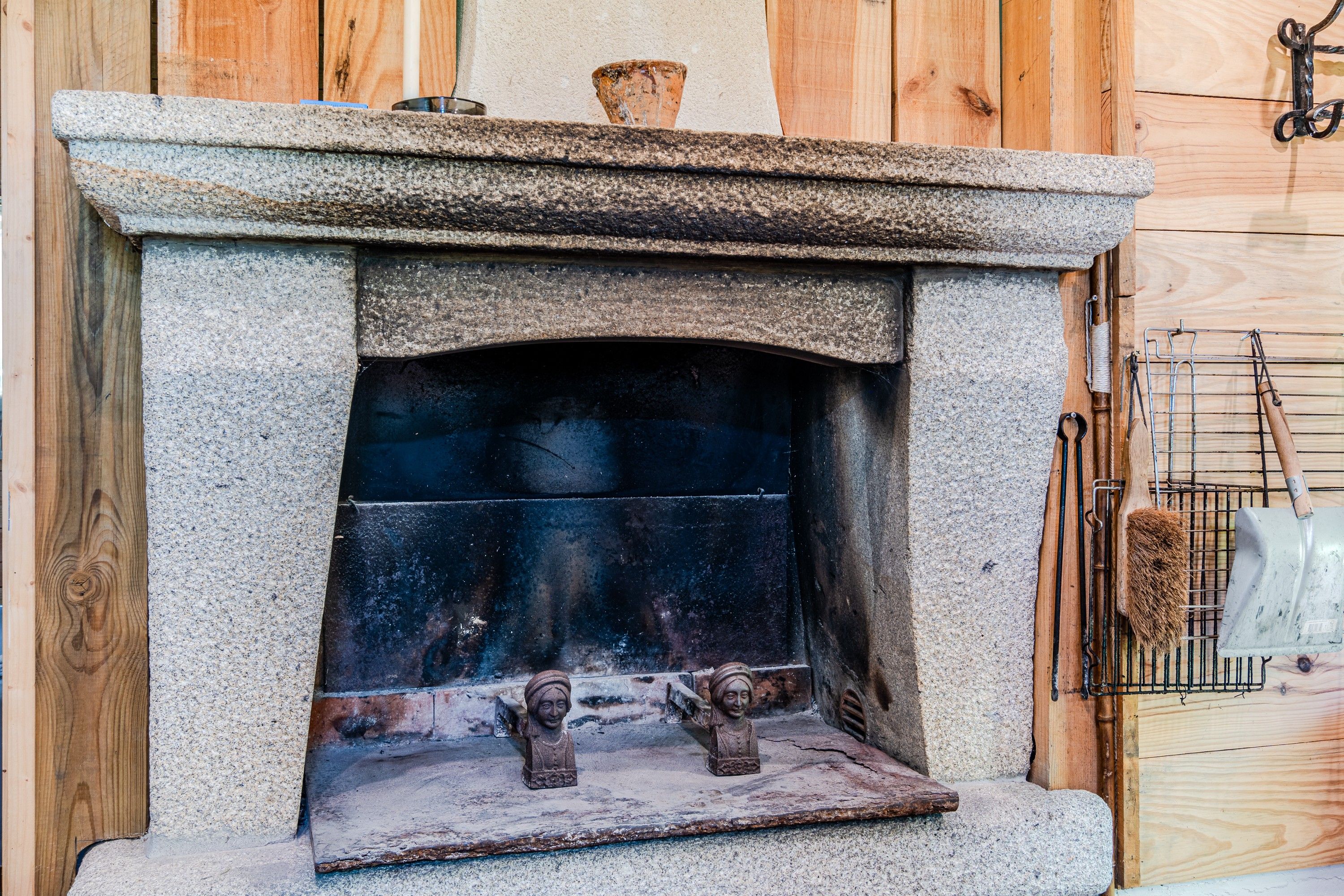 A stone fireplace with a metal grate, a rustic wooden wall, and a metal rack with tools.