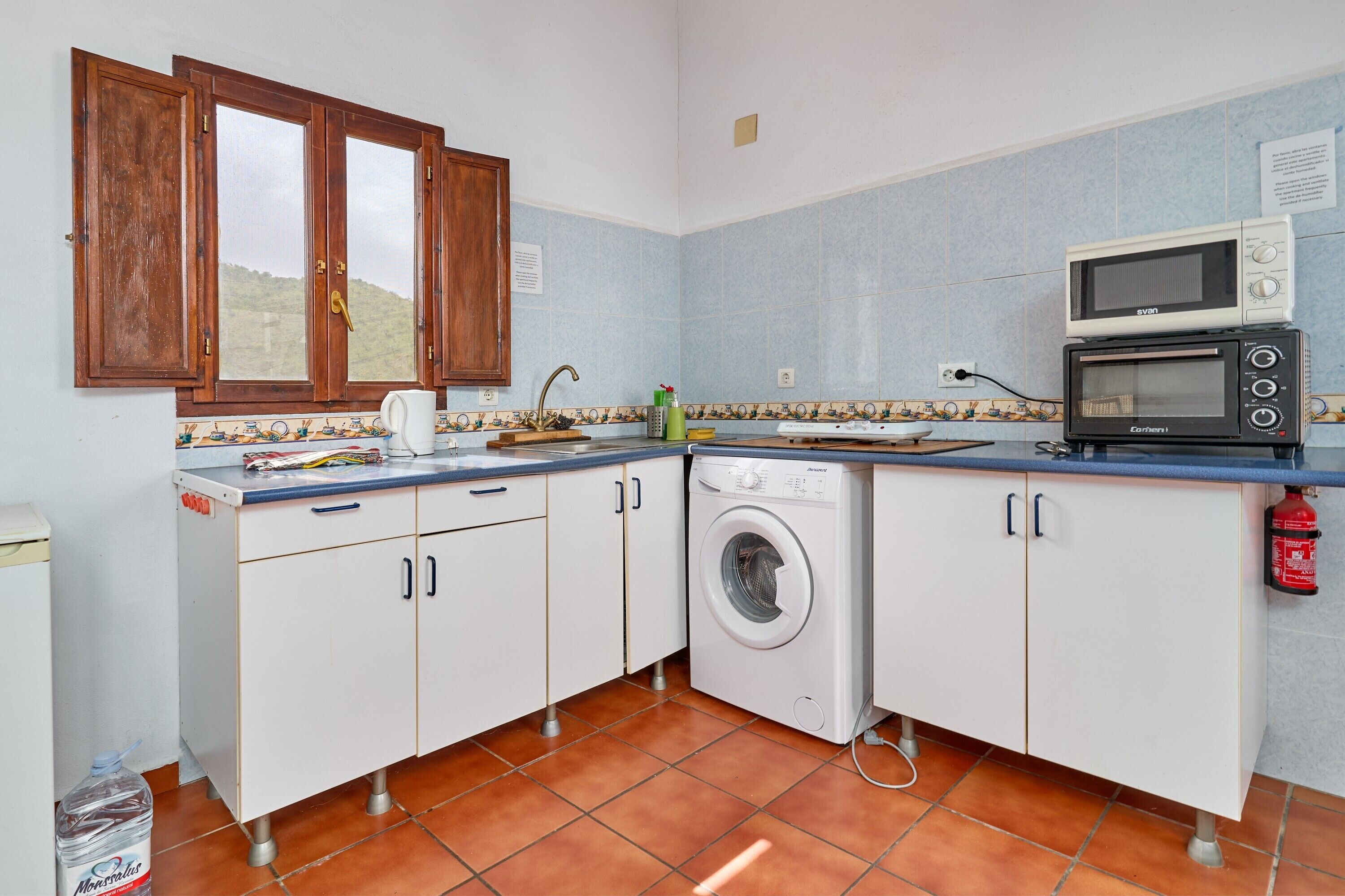 Une cuisine compacte équipée d’un lave-linge, d’un four à micro-ondes et d’une fenêtre avec des volets en bois.