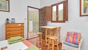 Una stanza con un letto, una cassettiera in legno, due sgabelli, una sedia in vimini con cuscino, un tavolino e una finestra con persiane di legno.