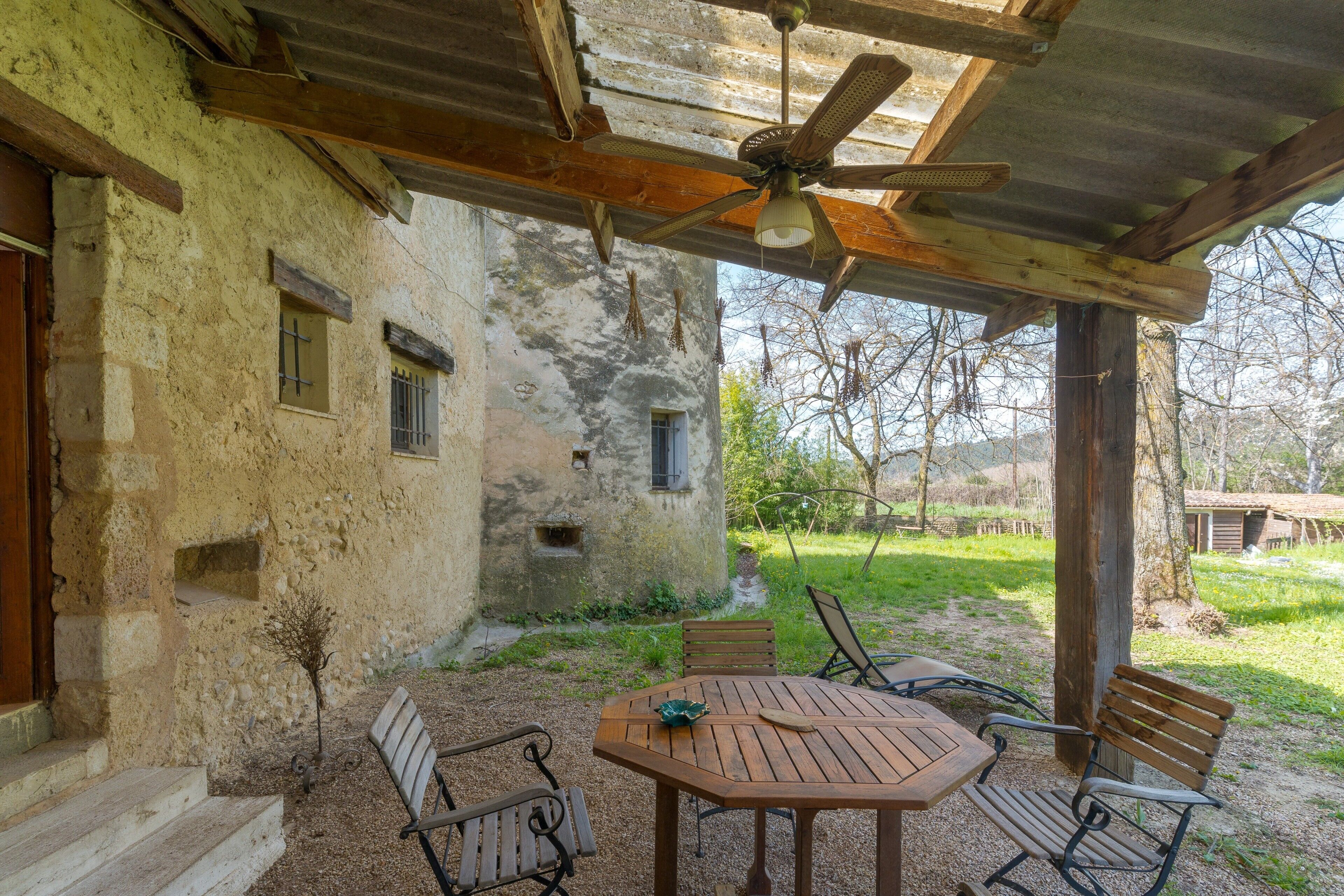 An outdoor patio with a wooden table and chairs, a ceiling fan, and a rustic building with a stone wall and wooden beams.