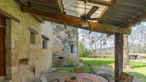 Un patio al aire libre con mesa y sillas de madera, un ventilador de techo y un edificio rústico con pared de piedra y vigas de madera.