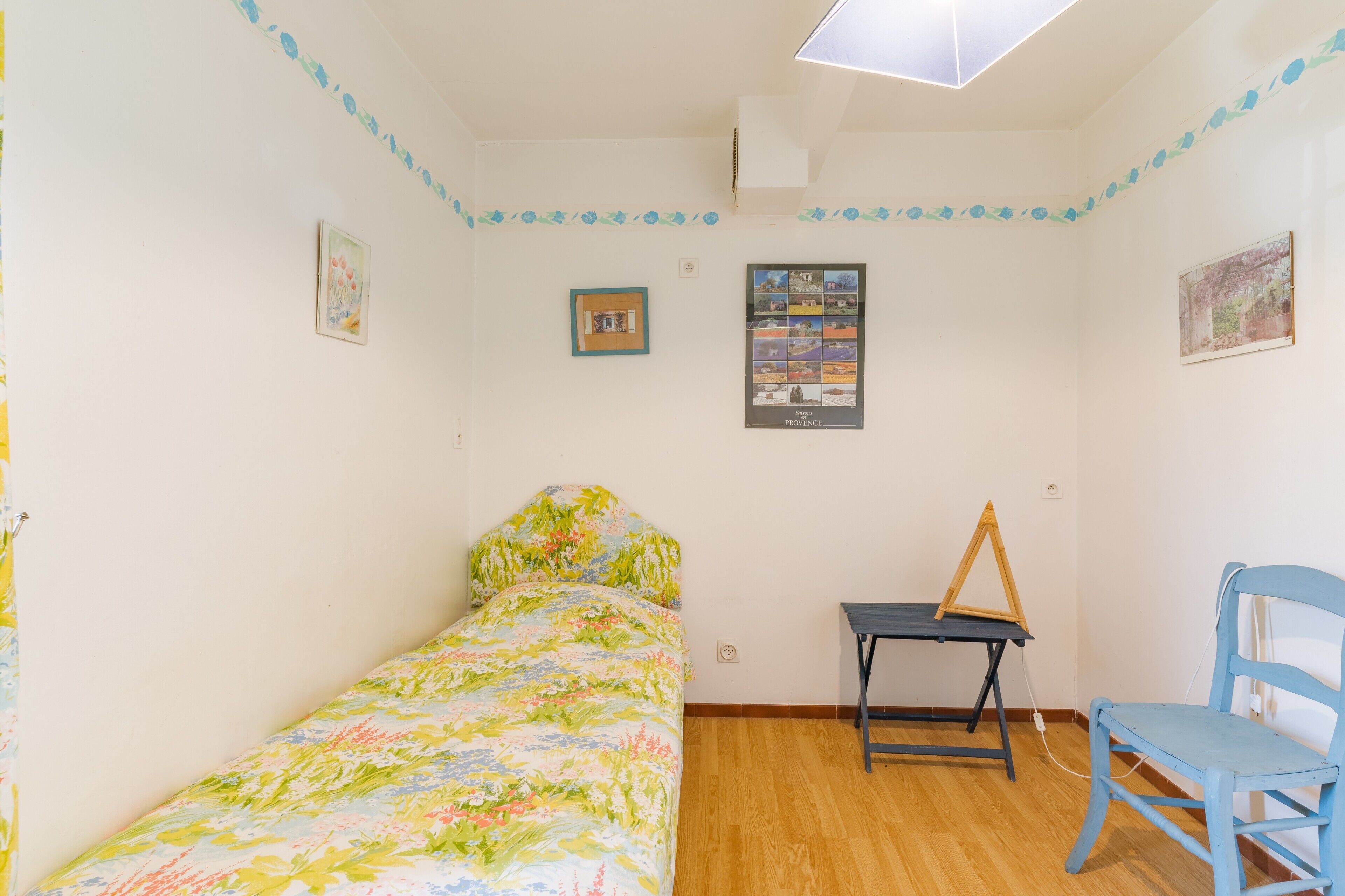 A small, single-bed room with a wooden floor, a desk, a chair, and wall decorations including framed pictures and a map.
