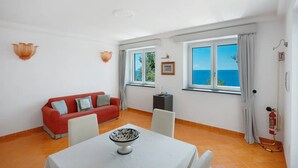 Una habitación con un sofá rojo, una mesa blanca con un bol, dos ventanas con cortinas y un extintor.