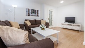 Ein modernes Wohnzimmer mit einer Sofagarnitur, einem Couchtisch mit Blumenvase, einem Flachbildfernseher auf einem weißen Schrank und einem Gemälde an der Wand.