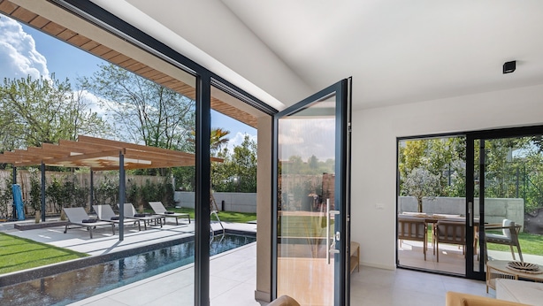 Ein modernes Haus mit Glastür zu einem Poolbereich, einer Terrasse mit Liegestühlen und einer Holz-Pergola.