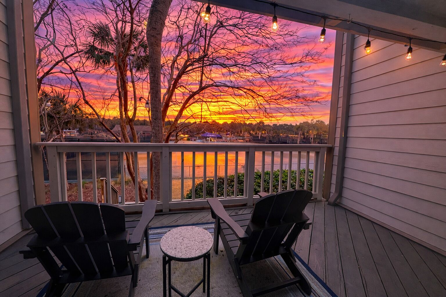 A covered porch with two chairs and a small table, overlooking a sunset over a body of water.