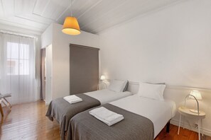 A hotel room with two beds, a wooden floor, a window with curtains, a door, and a lamp on a bedside table.
