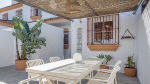 Un patio con una mesa y sillas blancas, plantas en macetas, un letrero direccional y una ventana con barrotes.