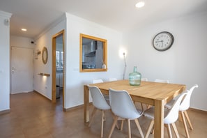 Un coin repas avec une table en bois, des chaises blanches, une horloge et une cuisine visible à travers une porte ouverte.