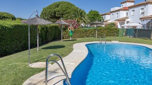 Una piscina con una escalera metálica, rodeada por un césped cuidado, una sombrilla de paja y un seto.