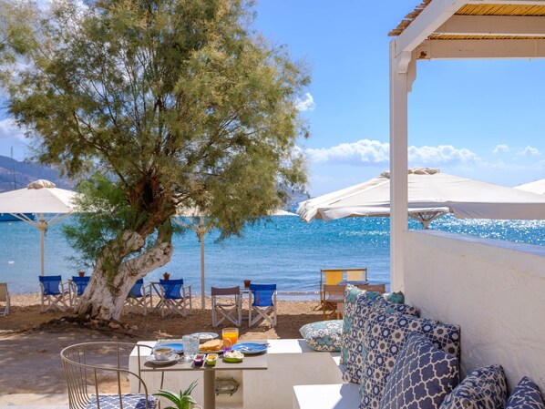 Une terrasse en bord de mer avec des sièges extérieurs, une table dressée pour le petit-déjeuner et des parasols pour faire de l’ombre.