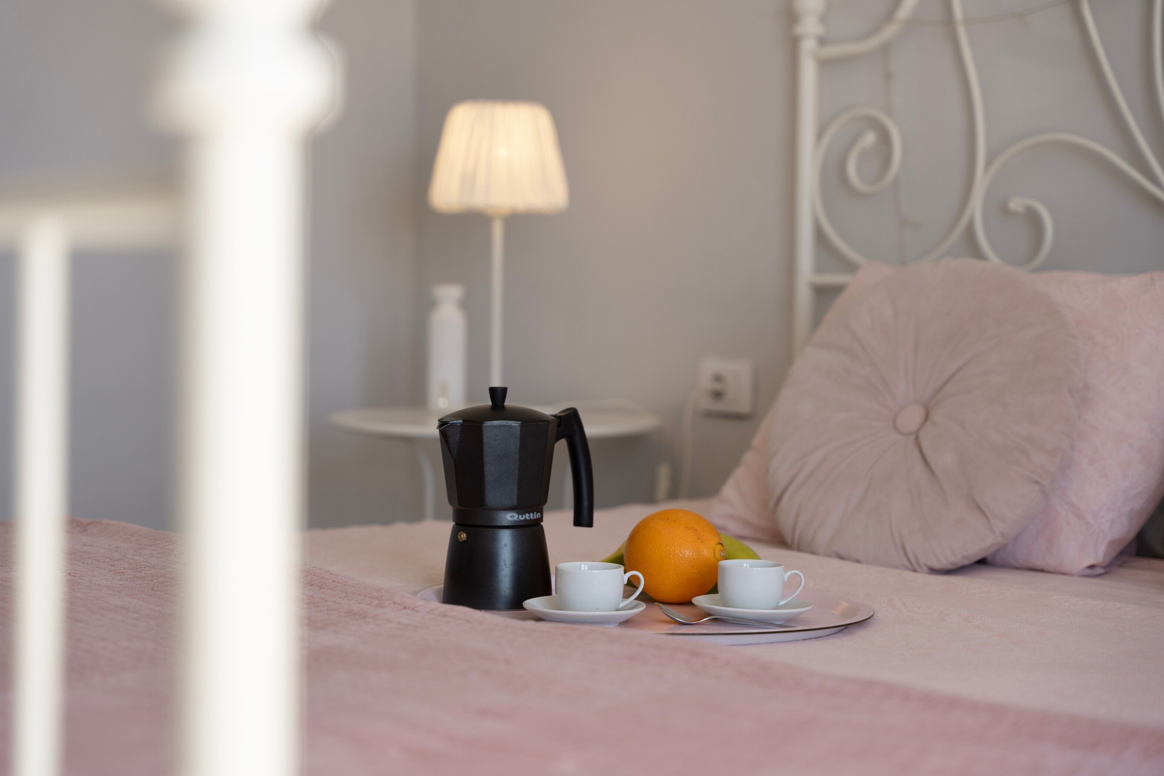 Un lit avec une couverture rose, un oreiller blanc, une machine à café, deux tasses blanches et une orange sur un plateau.