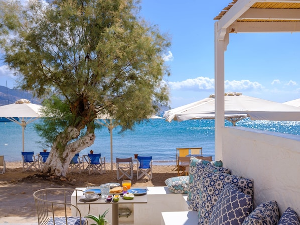 A seaside terrace with outdoor seating, a table set for breakfast, and umbrellas providing shade.