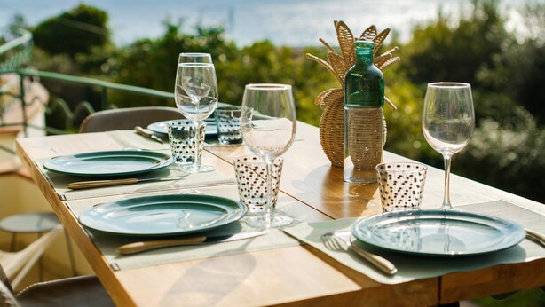A table set with plates, glasses, and cutlery, with a bottle and decorative basket on the table, overlooking a scenic view. - Mediterranean Escape Between the Sea and the Esterel Mountains (Théoule sur Mer)