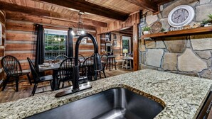 A rustic interior with a stone fireplace, wooden walls, a granite countertop, and a dining area with black chairs and a hanging light fixture.