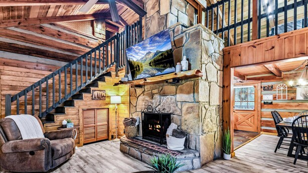 A log cabin interior with a stone fireplace, wooden staircase, leather sofa, and a dining area.