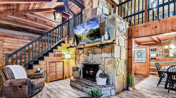 A log cabin interior with a stone fireplace, wooden staircase, leather sofa, and a dining area.
