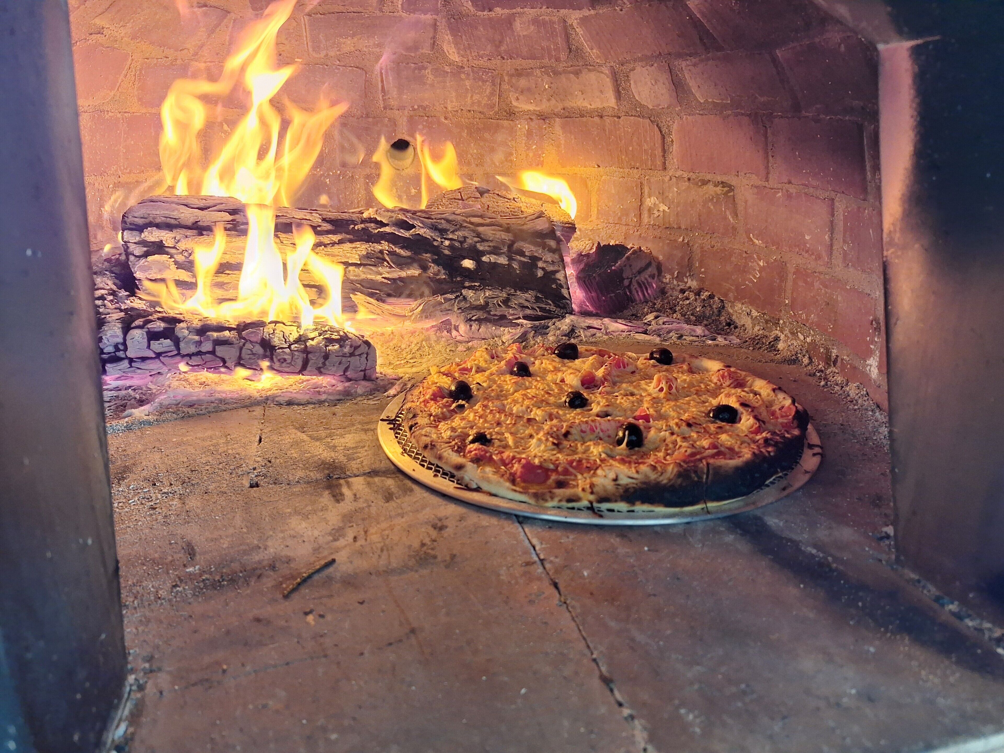 A pizza oven with a pizza inside, flames burning logs, and a brick wall.