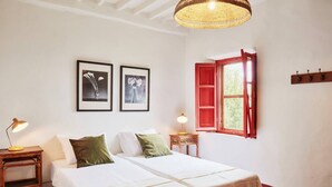 Un dormitorio con dos camas, una lámpara colgante, un ventanal con contraventanas rojas y ganchos en la pared.