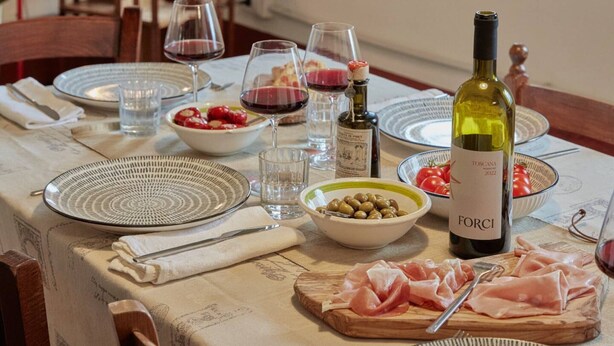 Una mesa de comedor servida con platos, vasos, una botella de vino y comida.