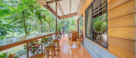 A covered outdoor seating area with wooden furniture, a view of lush greenery, and a large window.