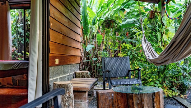 A wooden exterior wall, a hammock, a black chair, and a small table with a glass top. - Colibri Cabin (La Fortuna)