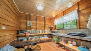 A wooden interior with a round dining table, bar stools, a kitchen area with appliances, and a window with a view. - Colibri Cabin (La Fortuna)