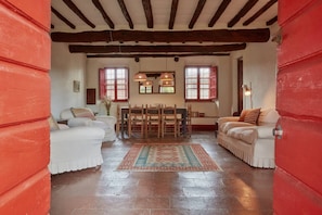 A room with a wooden ceiling, a dining table with chairs, a sofa, and a patterned rug.