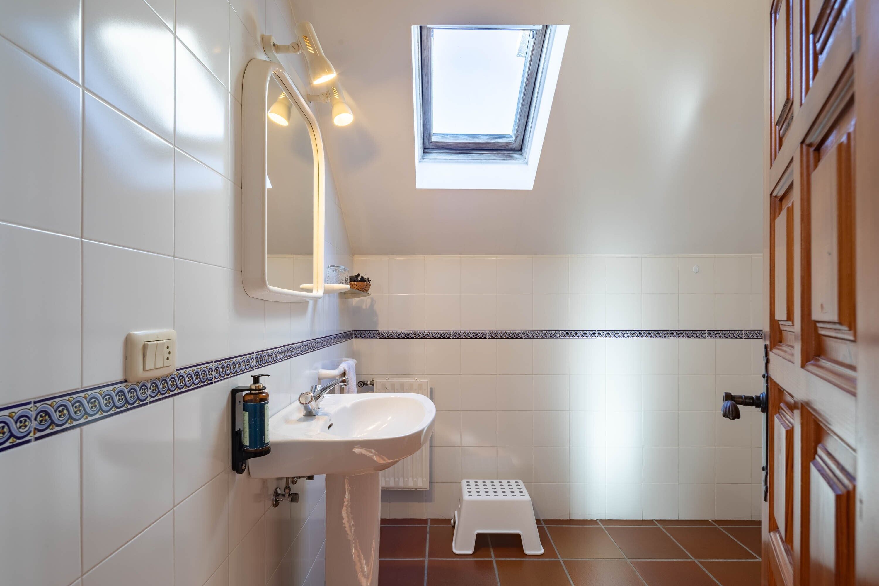 Un baño con tragaluz, azulejos blancos, un lavabo y un taburete pequeño.