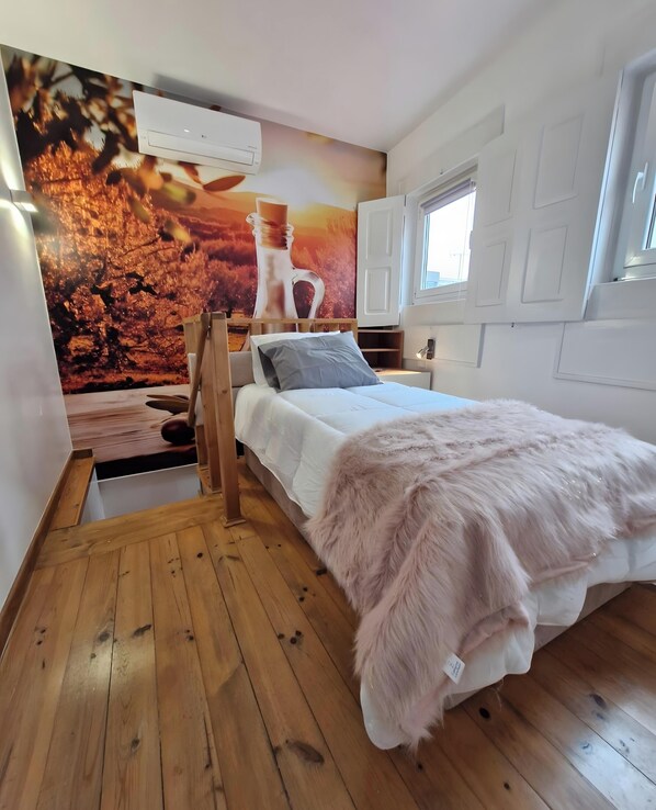 Une chambre à coucher avec un lit, un plancher en bois, un mur blanc et une fenêtre.