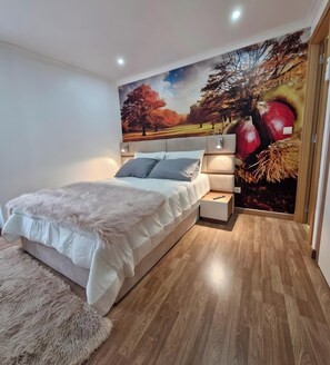 Une chambre à coucher comprenant un lit, une table de chevet, une lampe fixée au mur et une fresque représentant des arbres d’automne.
