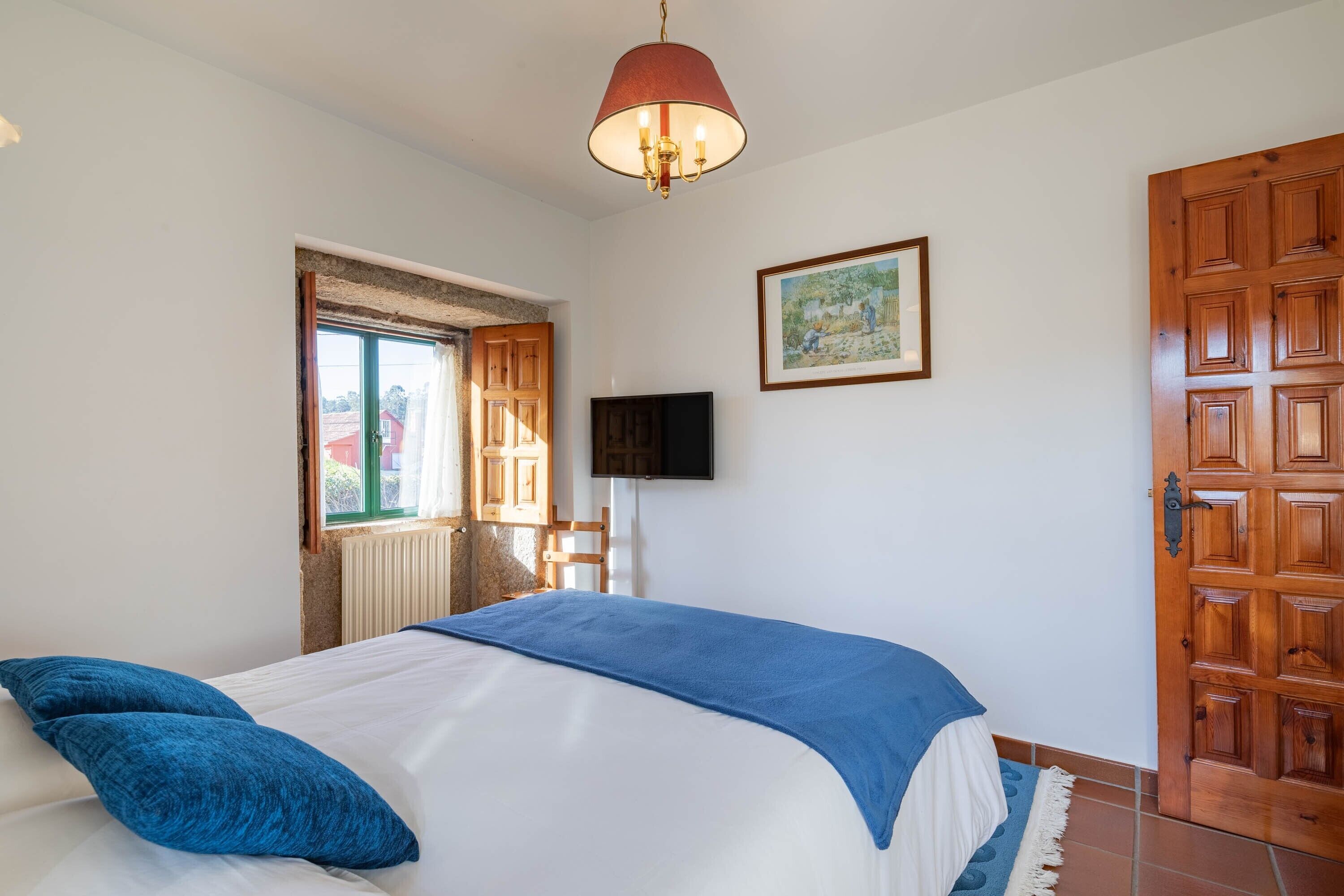 Un dormitorio con una cama, televisor, puerta de madera y ventana con cortinas.