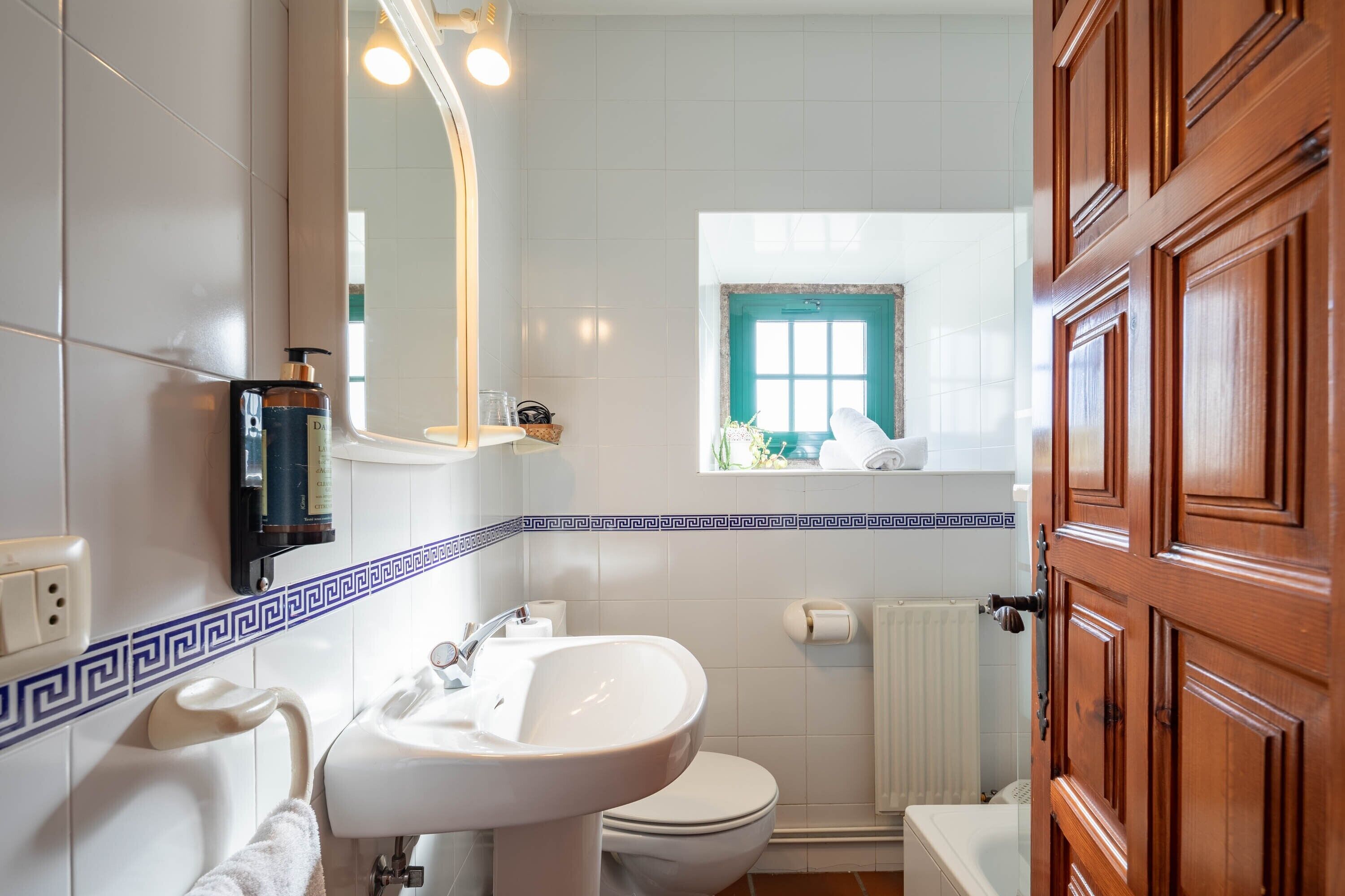 A bathroom with a white sink, toilet, and mirror, a wooden door, and a window with a view of the outside.