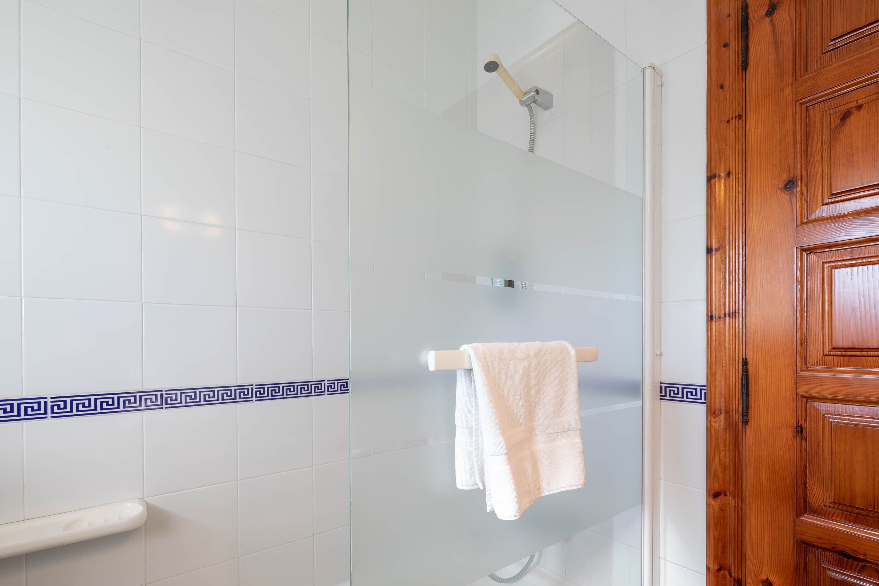 Un baño con paredes de azulejos blancos, una ducha con puerta de vidrio y una puerta de madera.